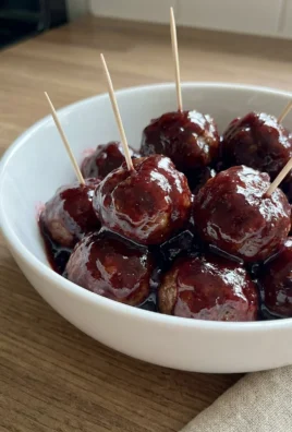 A white ceramic bowl filled with glossy grape jelly meatballs with toothpicks in them.