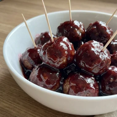 A white ceramic bowl filled with glossy grape jelly meatballs with toothpicks in them.